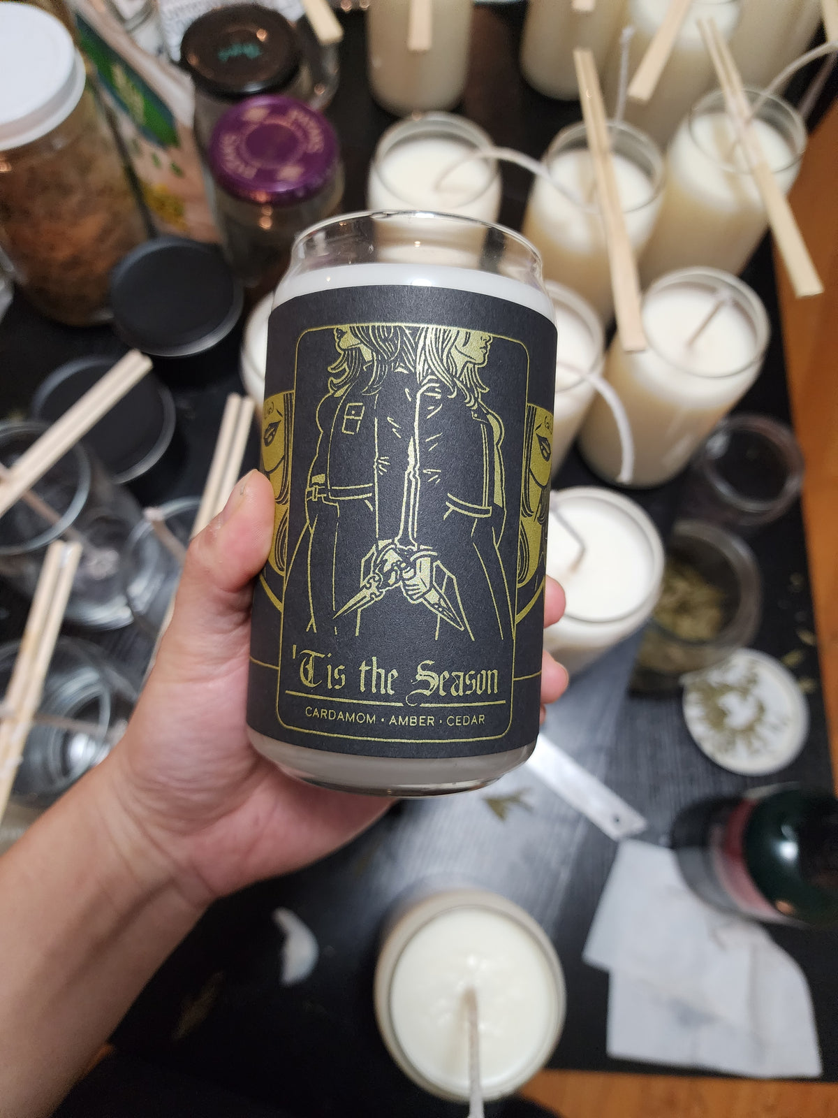 A hand holds a Buffering the Vampire Slayer candle labeled "Tis the Season: Cardamom · Amber · Cedar," featuring two people in winter coats. Other candles, supplies, and cedar sprigs are visible on the table in the background.