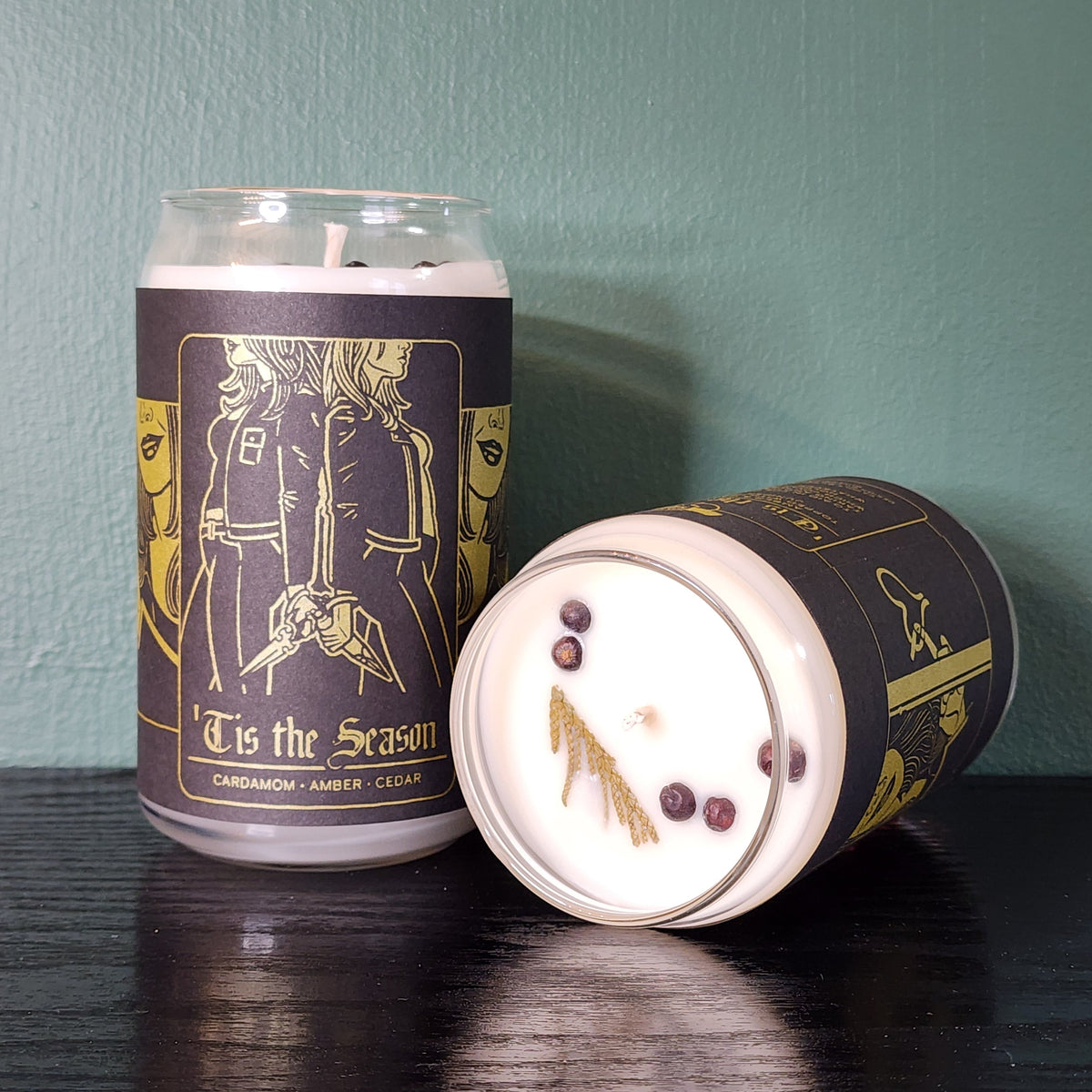Two handmade white candles by Buffering the Vampire Slayer in glass jars—one upright, one lying down—with dark “‘Tis the Season” labels featuring gold art of two people holding hands. Cedar sprigs top each candle.