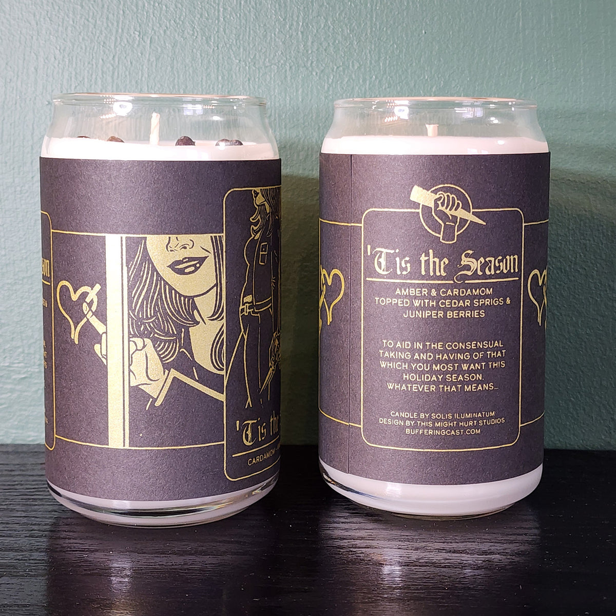 From Buffering the Vampire Slayer: Two white handmade candles in glass jars, featuring black and gold labels with illustrated hands, blending amber, cardamom, cedar sprigs, and juniper berries for a festive scent.