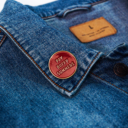 A close-up of a blue denim jacket with a round red enamel pin on the collar from Buffering the Vampire Slayer that reads PAY BUFFY SUMMERS in gold letters.