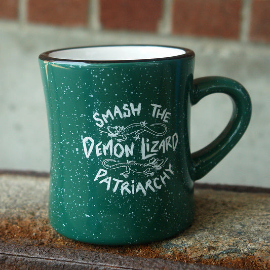 A green speckled mug from Buffering the Vampire Slayer, designed by Isabella Rotman, features “Smash the Demon Lizard Patriarchy” in white text and two cartoon lizards. Its photographed on a rough surface with a brick wall behind it.