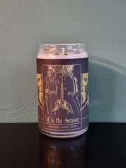 A handmade candle from Buffering the Vampire Slayer in a can features a label with two women back to back, holding hands. The label reads "Tis the Season" and highlights cardamom, amber, and cedar scents. It sits on a dark surface against a green wall.