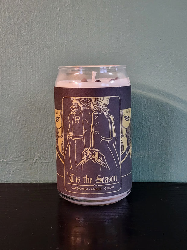 A handmade candle from Buffering the Vampire Slayer in a can features a label with two women back to back, holding hands. The label reads "Tis the Season" and highlights cardamom, amber, and cedar scents. It sits on a dark surface against a green wall.