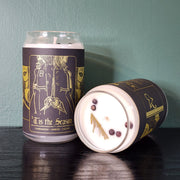 Two handmade white candles by Buffering the Vampire Slayer in glass jars—one upright, one lying down—with dark “‘Tis the Season” labels featuring gold art of two people holding hands. Cedar sprigs top each candle.