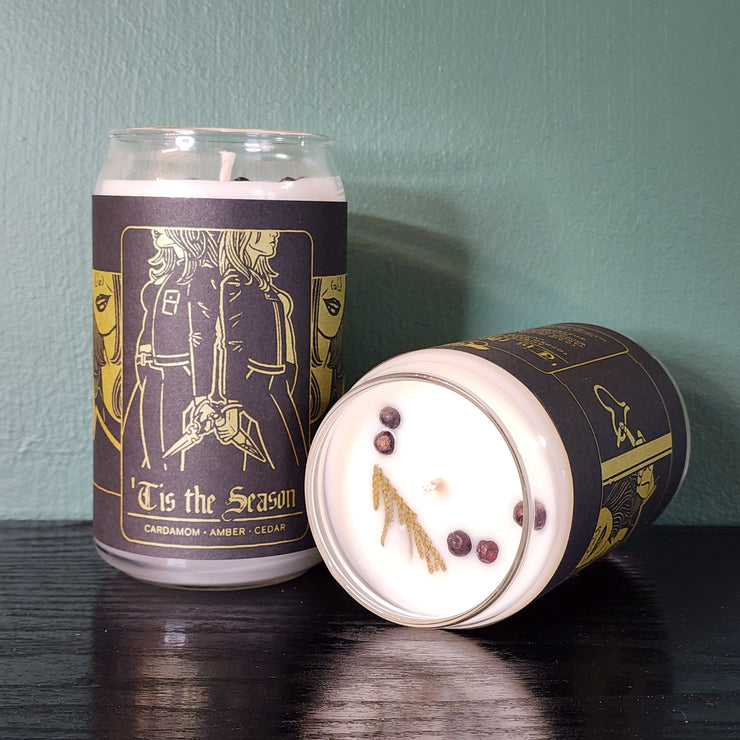 Two handmade white candles by Buffering the Vampire Slayer in glass jars—one upright, one lying down—with dark “‘Tis the Season” labels featuring gold art of two people holding hands. Cedar sprigs top each candle.