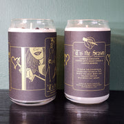 From Buffering the Vampire Slayer: Two white handmade candles in glass jars, featuring black and gold labels with illustrated hands, blending amber, cardamom, cedar sprigs, and juniper berries for a festive scent.
