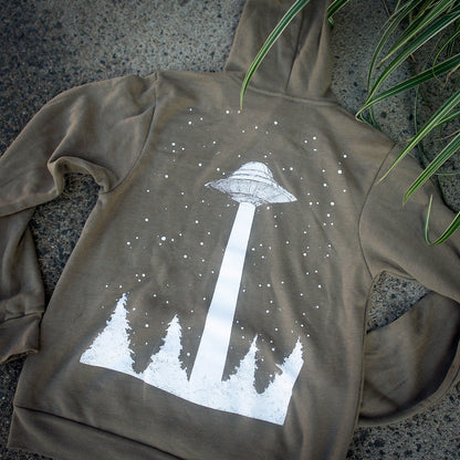 photo of the Want To Believe Heather - Olive Pullover. back has white print of a UFO with a beam down to a tree line forest laid flat outside