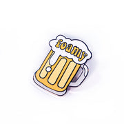 A quirky enamel pin from Buffering the Vampire Slayer shaped like a yellow beer mug with white foam, "foamy" in yellow letters across the suds, and sized like a 1.5" label for a playful accessory.