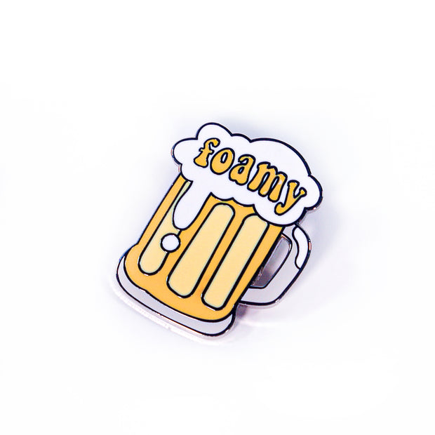 A quirky enamel pin from Buffering the Vampire Slayer shaped like a yellow beer mug with white foam, "foamy" in yellow letters across the suds, and sized like a 1.5" label for a playful accessory.