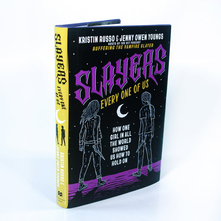A book titled Slayers: Every One of Us by Kristin Russo & Jenny Owen Youngs from Buffering the Vampire Slayer stands upright. The Buffy-inspired cover features two girls facing away beneath a crescent moon and stars in purple, yellow, and black.