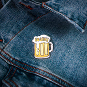 A foamy beer mug pin from Buffering the Vampire Slayer, featuring white foam and yellow liquid, is attached to blue denim fabric next to a small adhesive label for added flair.