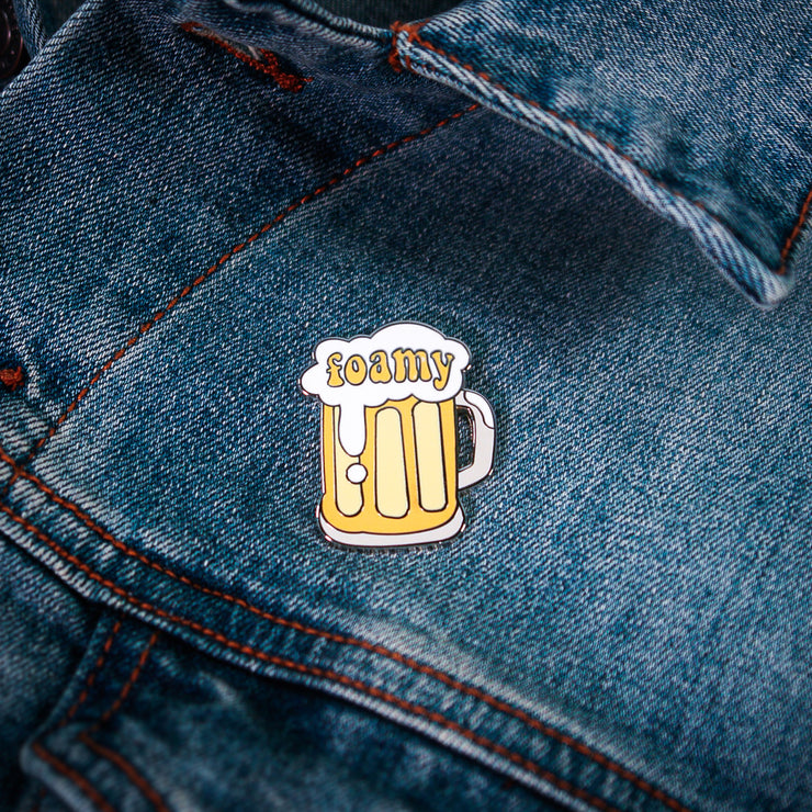 A foamy beer mug pin from Buffering the Vampire Slayer, featuring white foam and yellow liquid, is attached to blue denim fabric next to a small adhesive label for added flair.