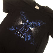 image of a black tee shirt on a white background. tee has center chest print of a giant letter X, with a man and woman facing away from each other holding flashlights