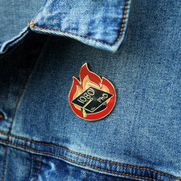 image of a enamel pin, pinned on a denim jacket. pin is in the shape of fire and has a graduation cap that says 1080 PhD