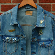 image of a hanging jean jacket in front of a brick building with enamel pins pinned on the jacket