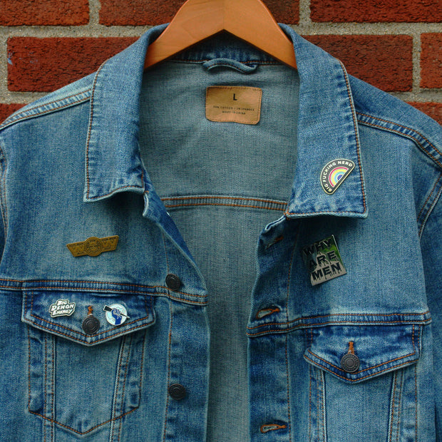 image of a hanging jean jacket in front of a brick building with enamel pins pinned on the jacket