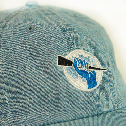 close up image of a denim dad hat on a white background. hat has small embroidery on the front of a circle, with a hand holding a stake