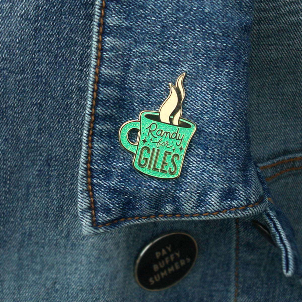 A turquoise enamel pin shaped like a steaming coffee cup with Randy for Giles by Buffering the Vampire Slayer is pinned to a blue denim jacket collar. Below it, there’s also a round black pin with white text.