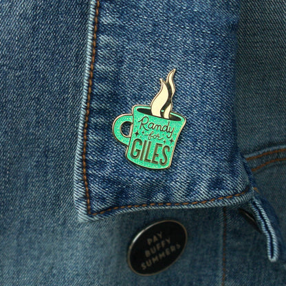 A turquoise enamel pin shaped like a steaming coffee cup with Randy for Giles by Buffering the Vampire Slayer is pinned to a blue denim jacket collar. Below it, there’s also a round black pin with white text.