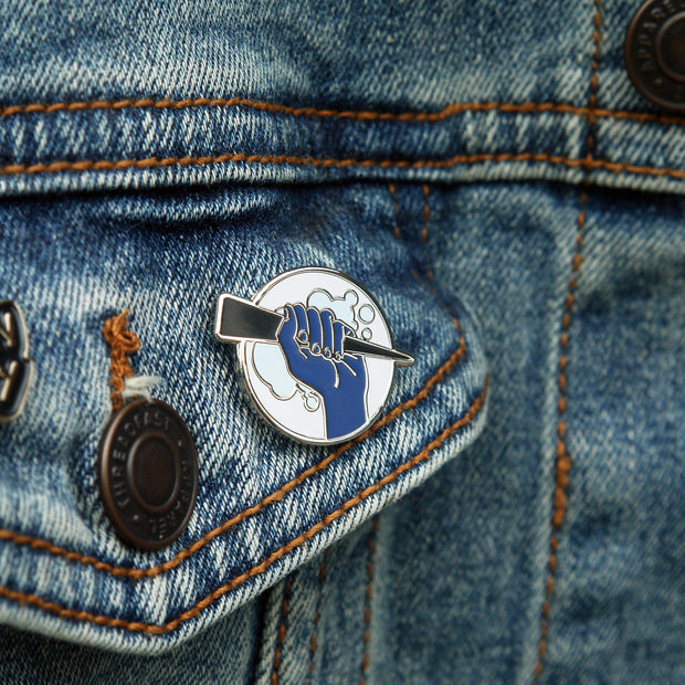 image of a circle enamel pin on a pinned on a jean jacket pocket. pin is of a hand holding a stake.