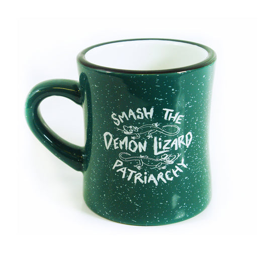 A dark green ceramic mug from Buffering the Vampire Slayer, designed by Isabella Rotman, features white speckles, small lizard illustrations, and the phrase Smash the Demon Lizard Patriarchy in white stylized text.