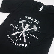 close up angled image of a black tee shirt on a white background. tee has center chest print in white that says smash the demon lizard patriarchy with two sledge hammers crossed and a stake in the middle